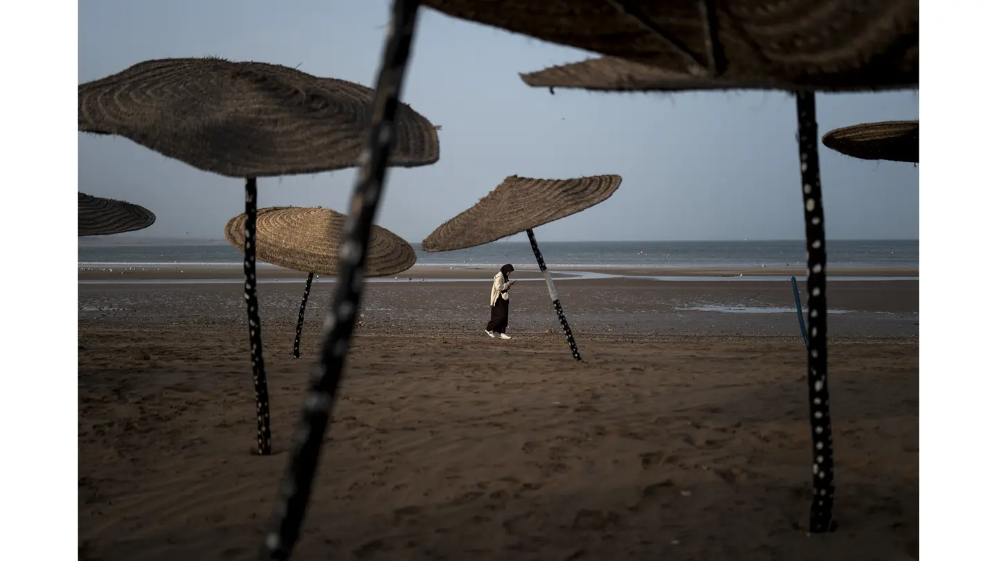 Une femme se promène sur une longue plage de sable, entourée de parasols en paille, tout en regardant son téléphone