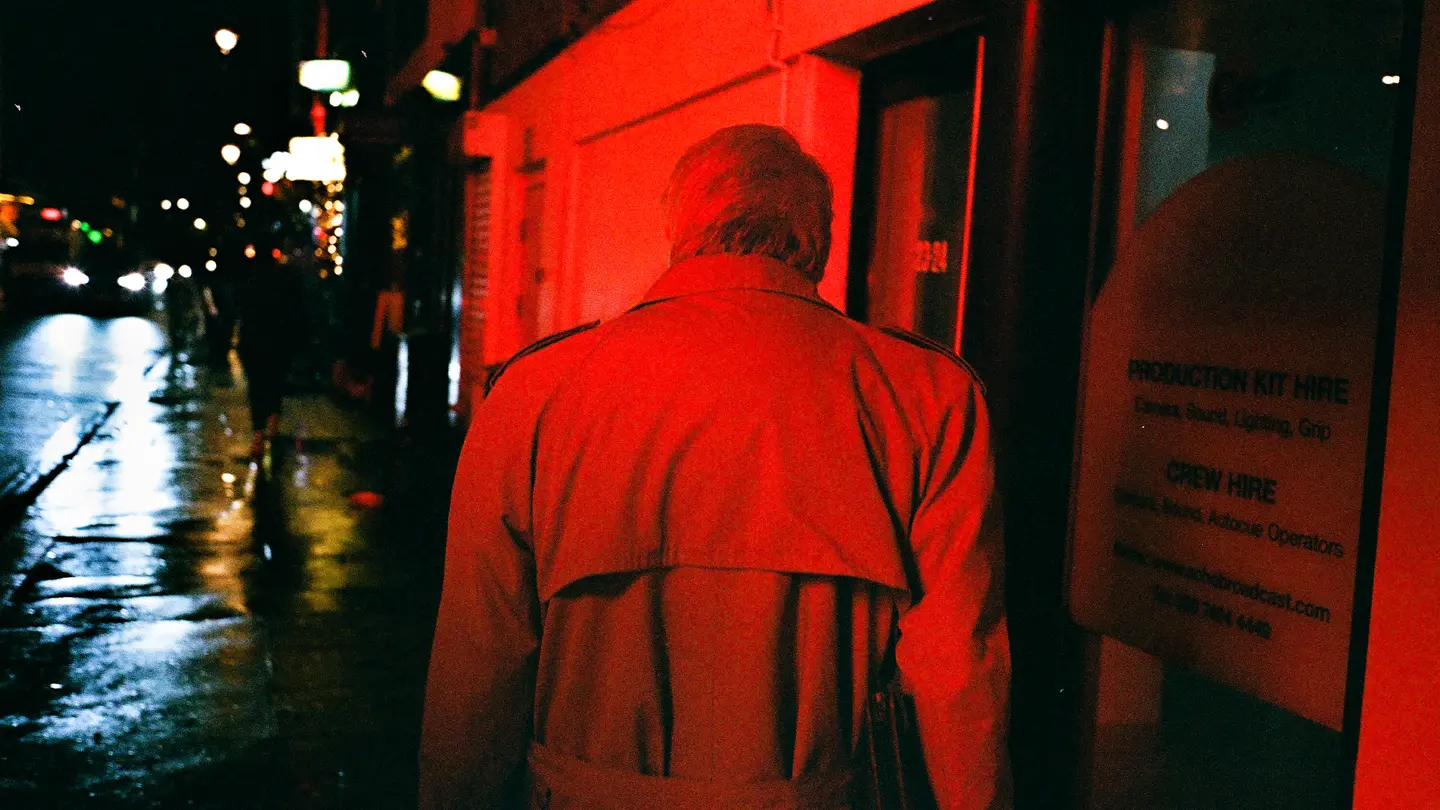 Colour photo of a man washed in red light walking on the highstreet at night.