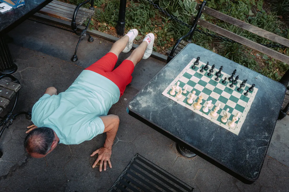 Person doing push-ups beside an outdoor chess table in a public park.