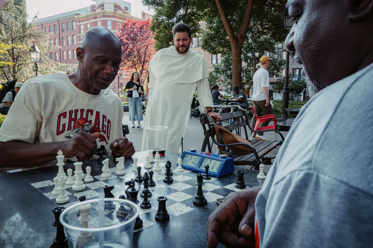 People playing chess in a public park while a passerby watches the game.