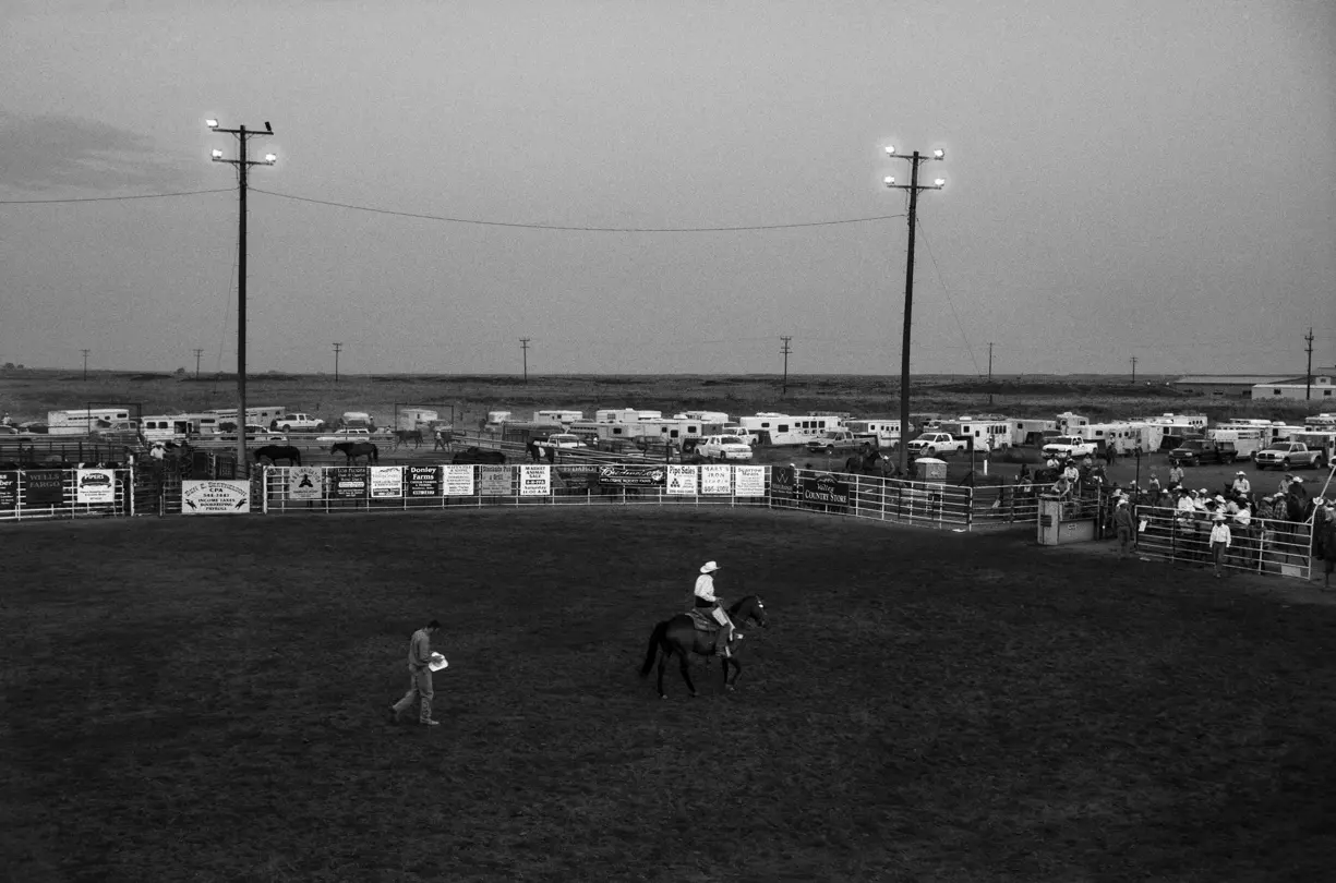 Black-and-white documentary photograph of a rodeo arena in rural Montana, where a rider on horseback prepares for competition during a local community event along Highway 93.