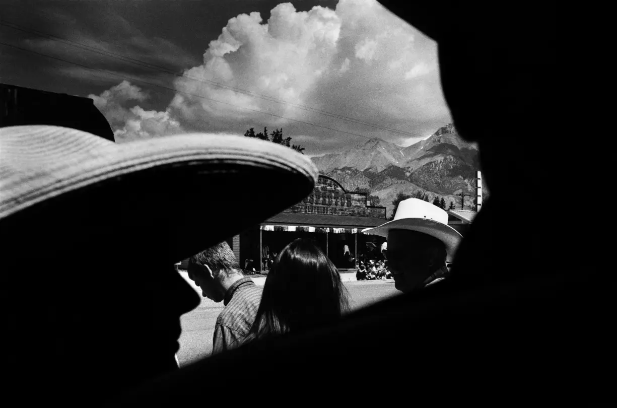 Black-and-white documentary photograph of people wearing cowboy hats in a small Montana town, with the Rocky Mountains rising in the background along Highway 93.