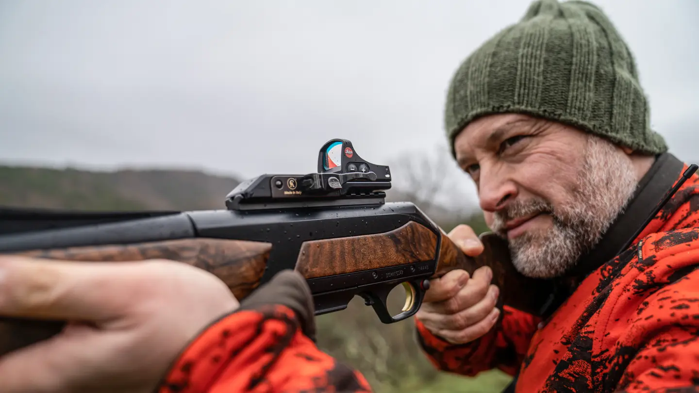 Close-up–of–a–hunter–aiming–a–rifle–with–a–Leica–red-dot–sight–while–wearing–orange–camouflage–and–a–green–beanie–outdoors
