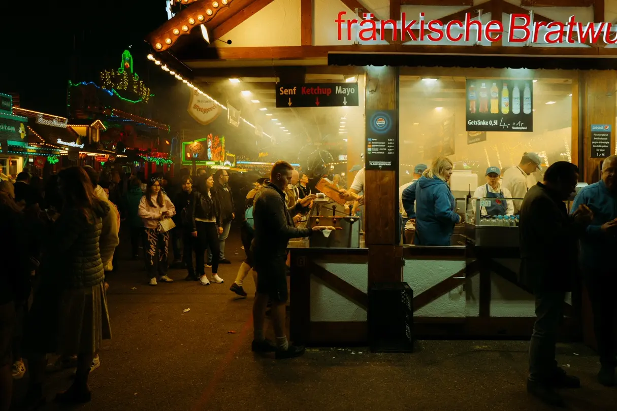 Busy bratwurst stand at night during Munich Oktoberfest, with waiting visitors and illuminated rides.