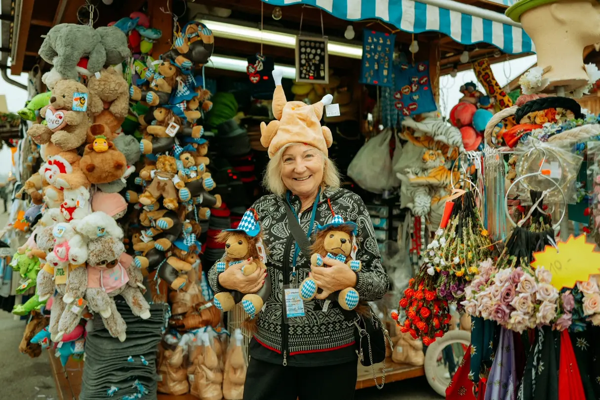 Smiling Oktoberfest vendor wearing a novelty chicken hat and holding Bavarian lion plush toys at a colorful souvenir stand in Munich.