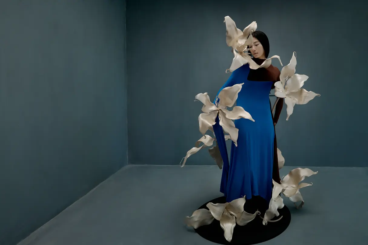 Model in blue dress — Woman in a vivid blue dress surrounded by sculptural white flowers in a minimalist blue-grey studio.