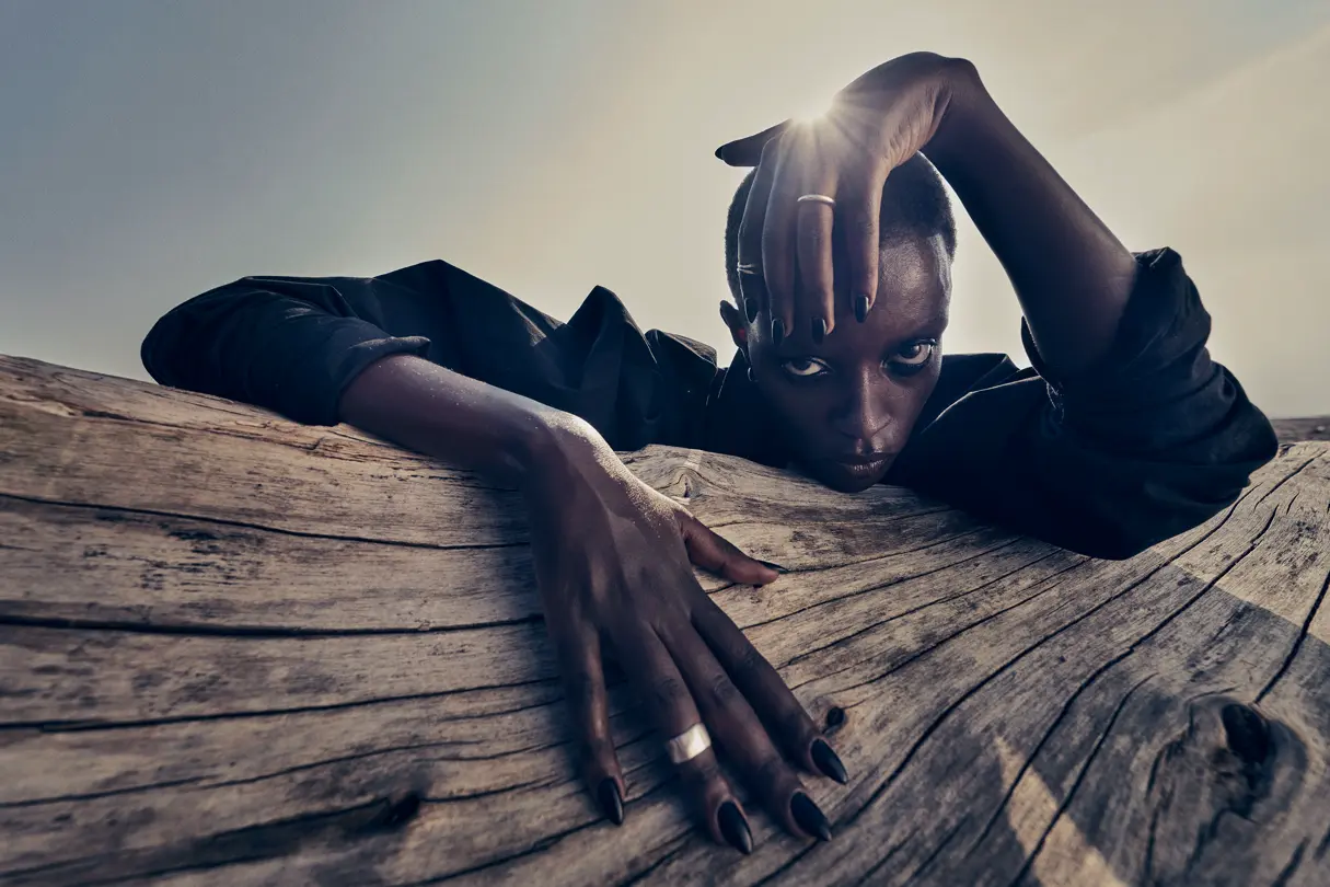 Dramatic fashion portrait outdoors — Low-angle portrait of a person in black leaning over weathered wood, with one hand framing their face as sunlight shines from behind.