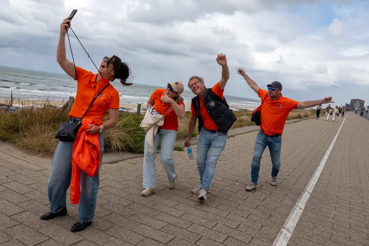 Group of Dutch Grand Prix fans in orange shirts cheer and take a selfie while walking along the seafront in Zandvoort.
