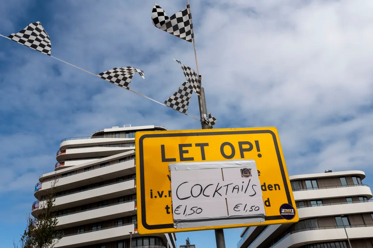 Checkered racing flags fly above a yellow warning sign with a handwritten “Cocktails” notice in Zandvoort during the Dutch Grand Prix.