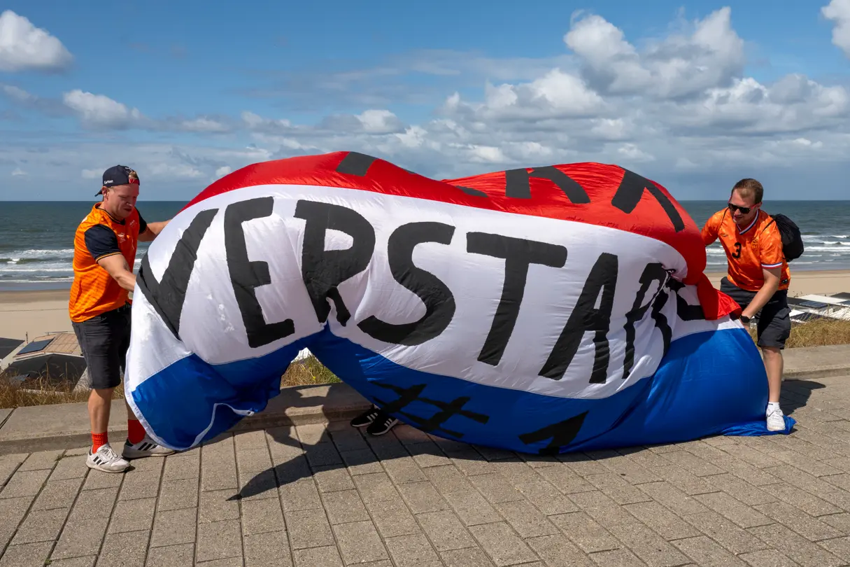 Two Formula 1 fans unfold a large red, white and blue Dutch flag with “Verstappen” lettering on the coast of Zandvoort.