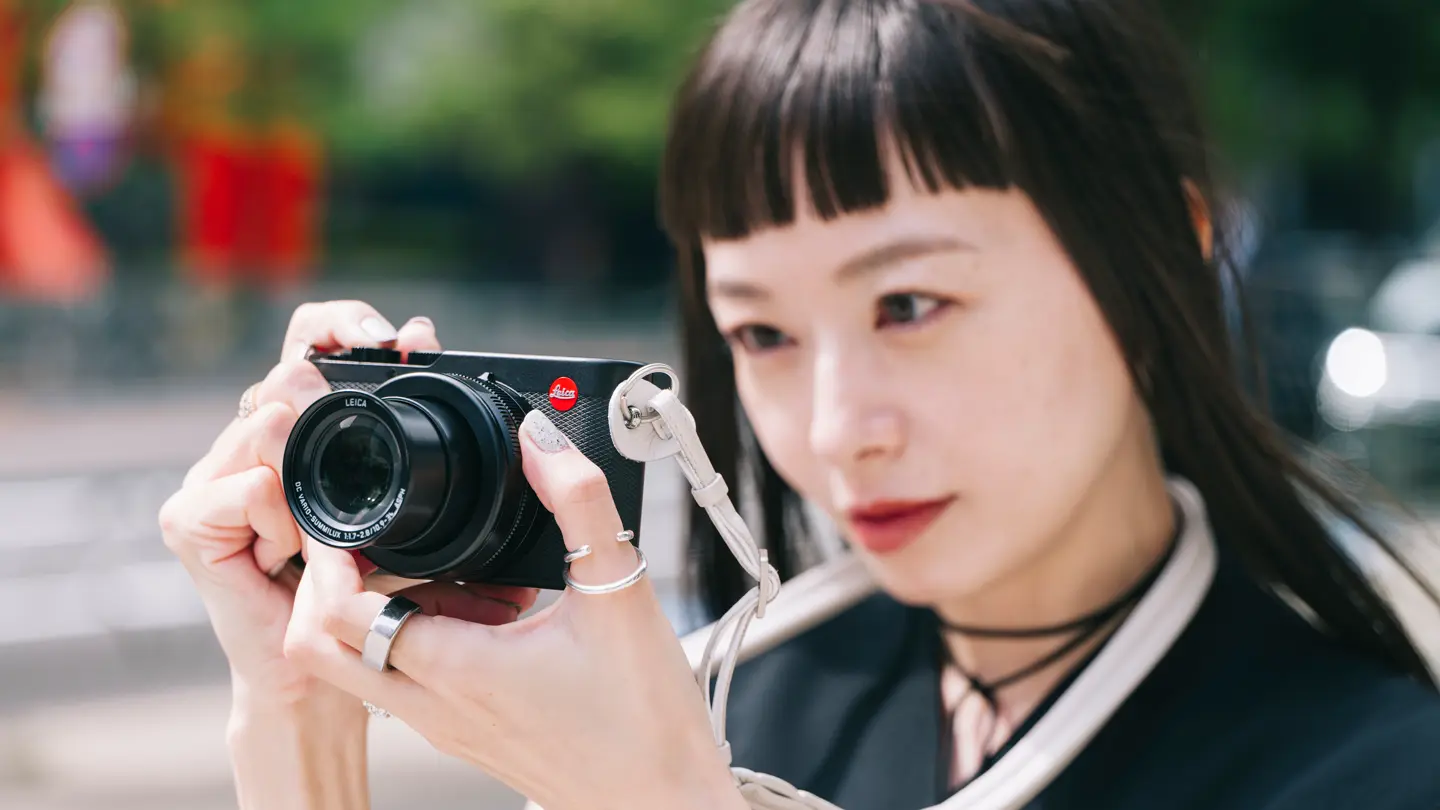 Woman using Leica camera — Young woman with dark hair holds a Leica compact camera up in front of her while photographing outdoors.