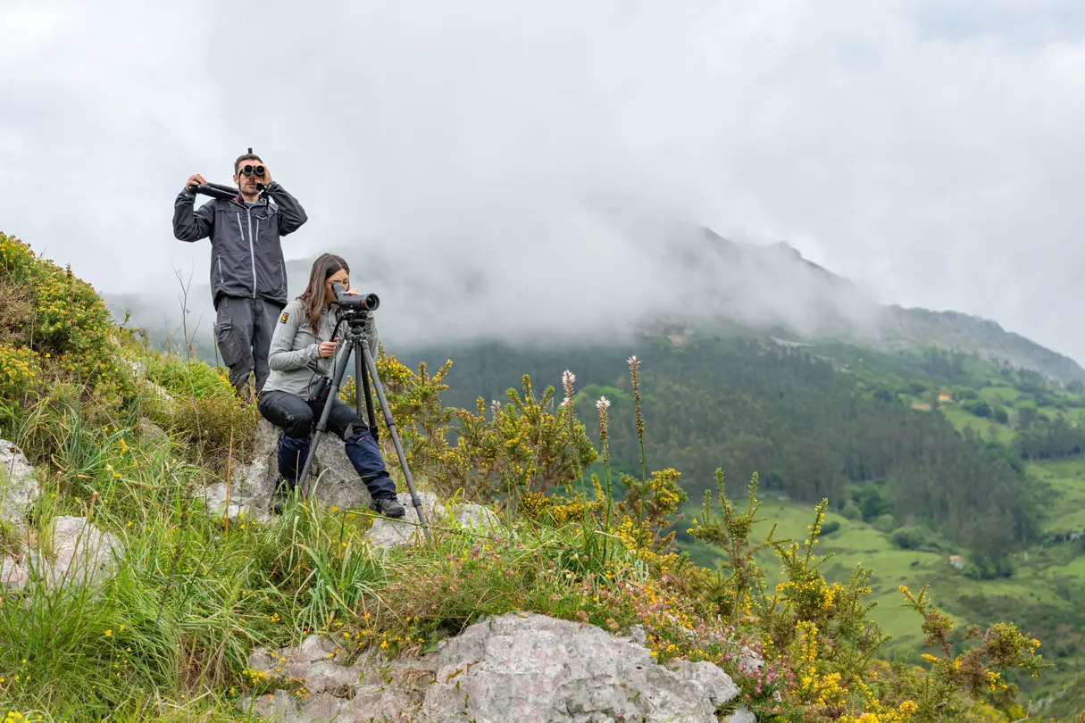 A man and a woman standing in the mountains and look through their binoculars