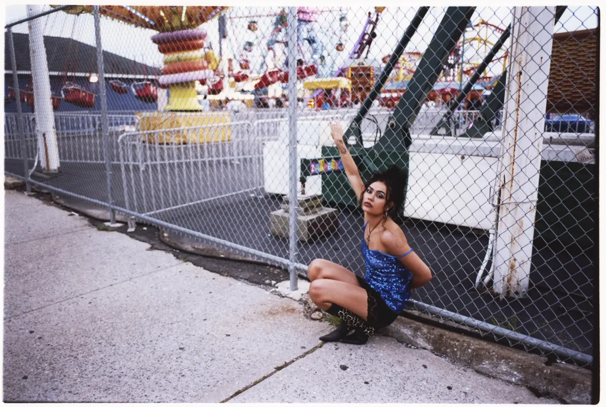 Coney Island: Woman in a blue sequined top poses beside a chain-link fence at an amusement park in Coney Island, New York.