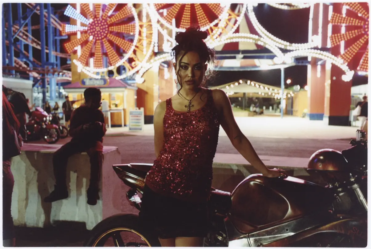 Coney Island at night: Woman in a red sequined top poses beside a motorcycle beneath illuminated amusement park rides at Coney Island, New York.