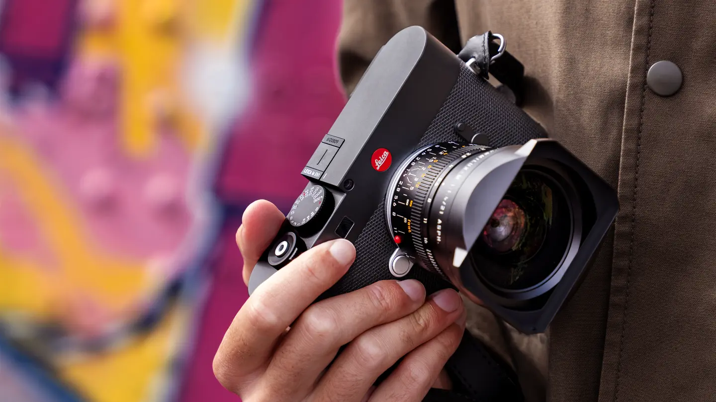 Leica M camera with wide-angle lens — Close-up of a black Leica M rangefinder camera with a wide-angle lens, held against a brown jacket with a colorful blurred background.