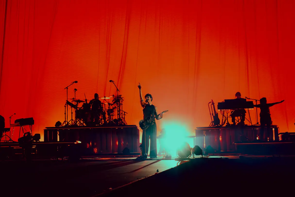 Live concert performance — Singer and guitarist performing on stage with a band, silhouetted against dramatic red and turquoise concert lighting.