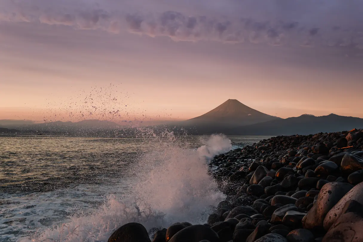 Mount Fuji at sunset — Waves crashing onto a rocky shoreline with Mount Fuji silhouetted beneath a soft pink and purple evening sky.