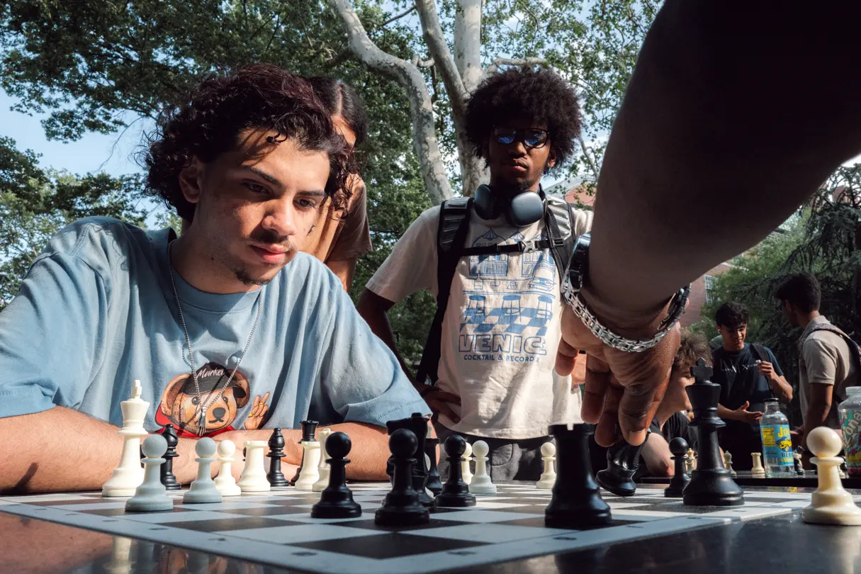 Outdoor chess game — Young people gathered around a chessboard in a park as a hand reaches in to move a chess piece.