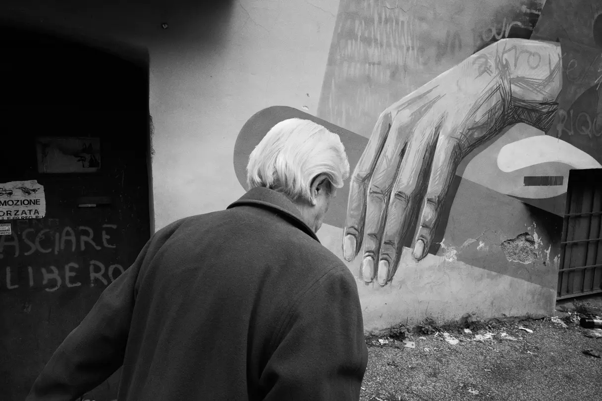 1. Black-and-white street photography — Elderly man walking past a large mural of a hand on a weathered urban wall in a documentary-style street photograph.