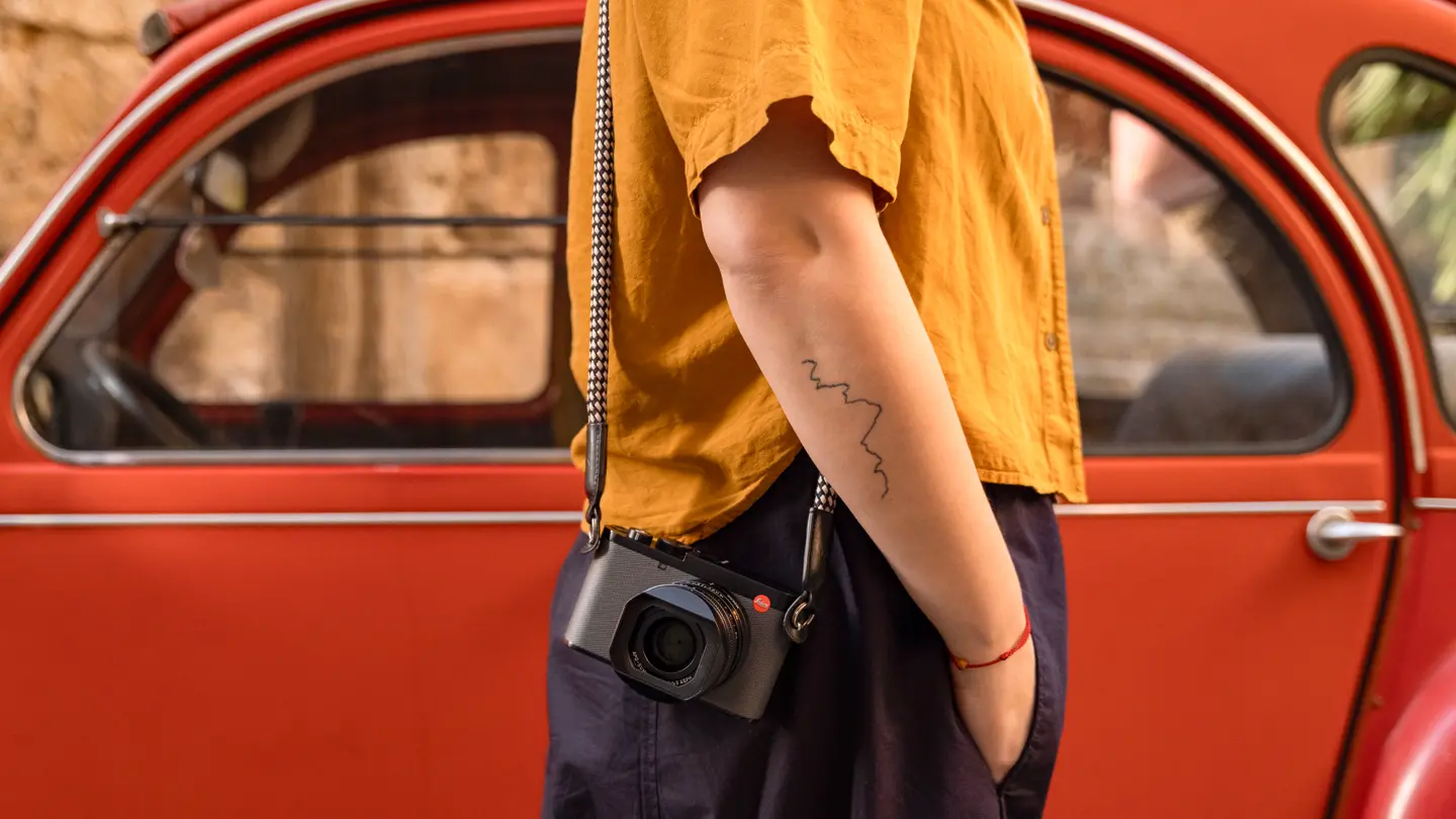 Person wearing a Leica Q compact camera on a rope strap while standing beside a classic red car, highlighting the camera’s compact design and travel-ready portability.