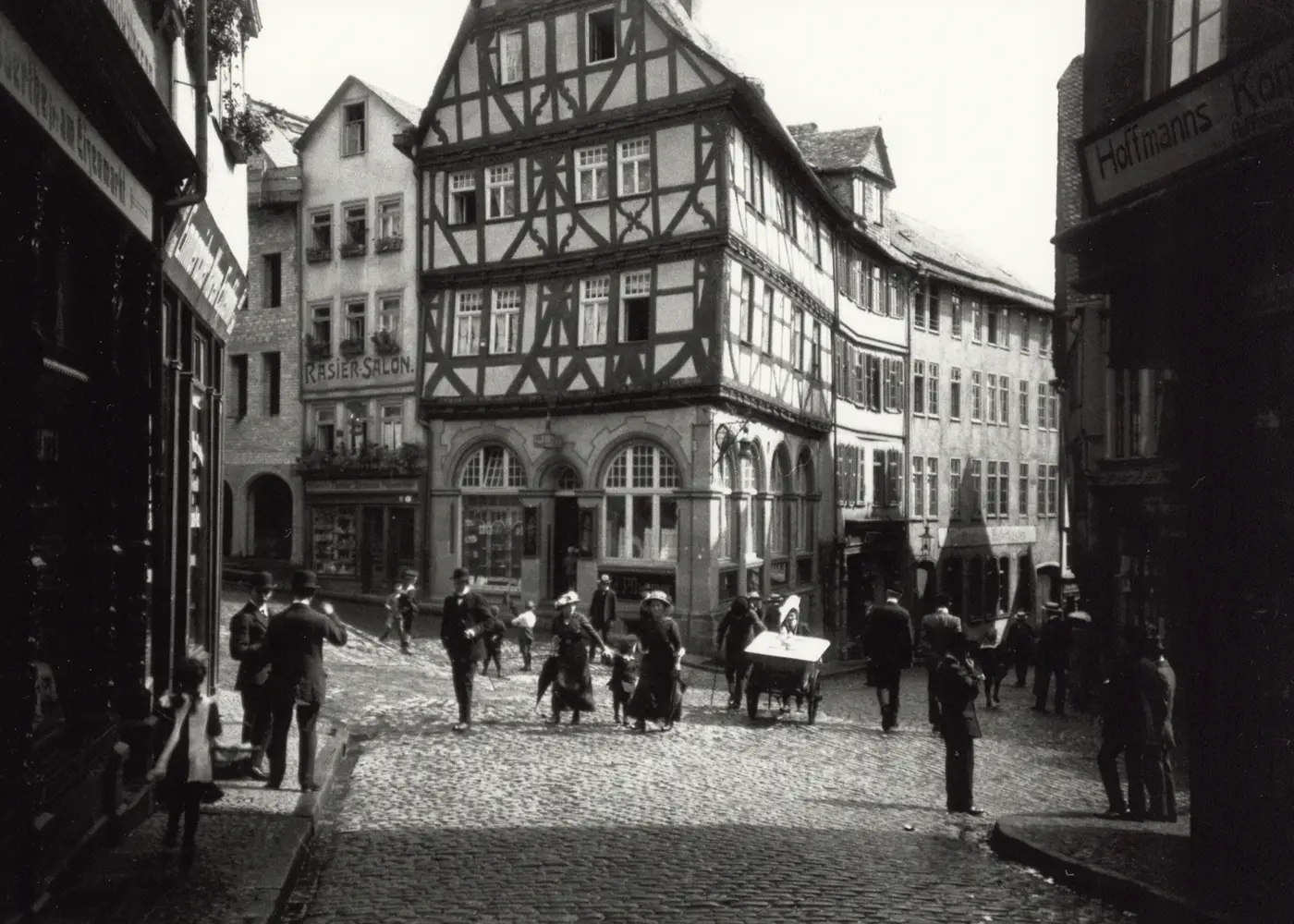 Historic black-and-white street scene with pedestrians walking past traditional half-timbered buildings on a cobblestone street in Germany.