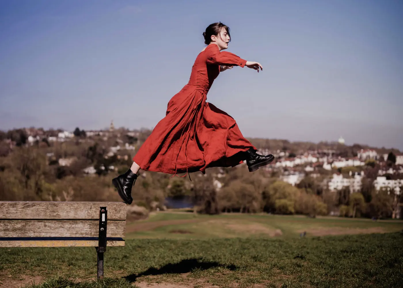 Woman in a flowing red dress captured mid-jump over a park bench, with a wide landscape and blue sky in the background.