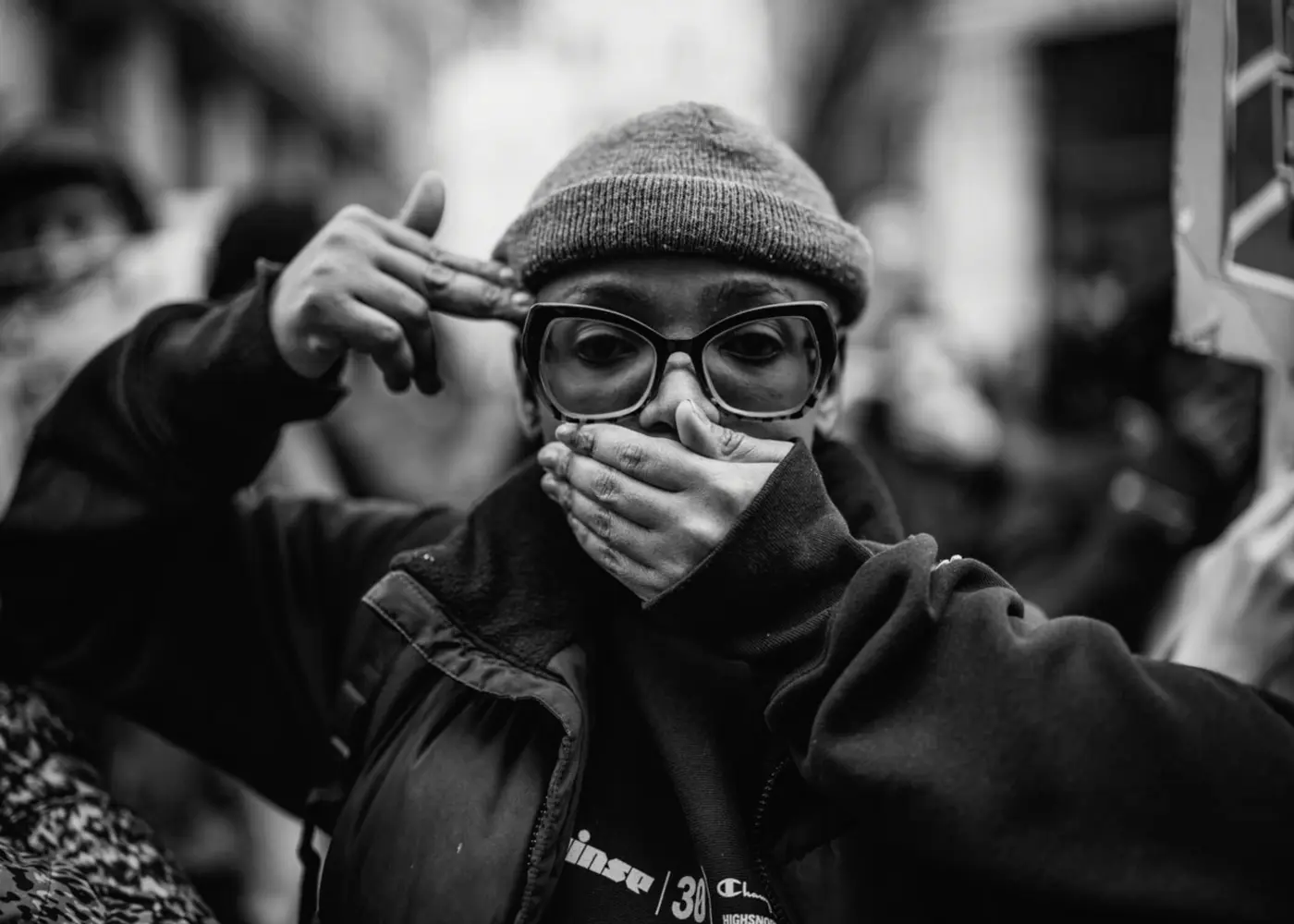 Black-and-white portrait of a person wearing a knit cap and oversized glasses, covering their mouth while another hand points toward their head.