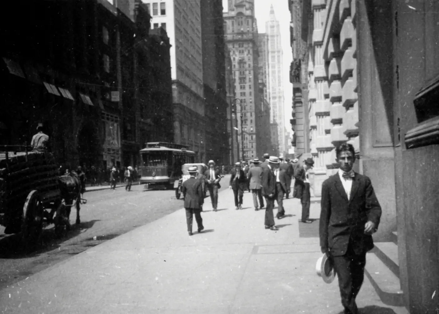 Historic black-and-white photograph of a busy city street with pedestrians, a streetcar and horse-drawn vehicles among tall buildings.