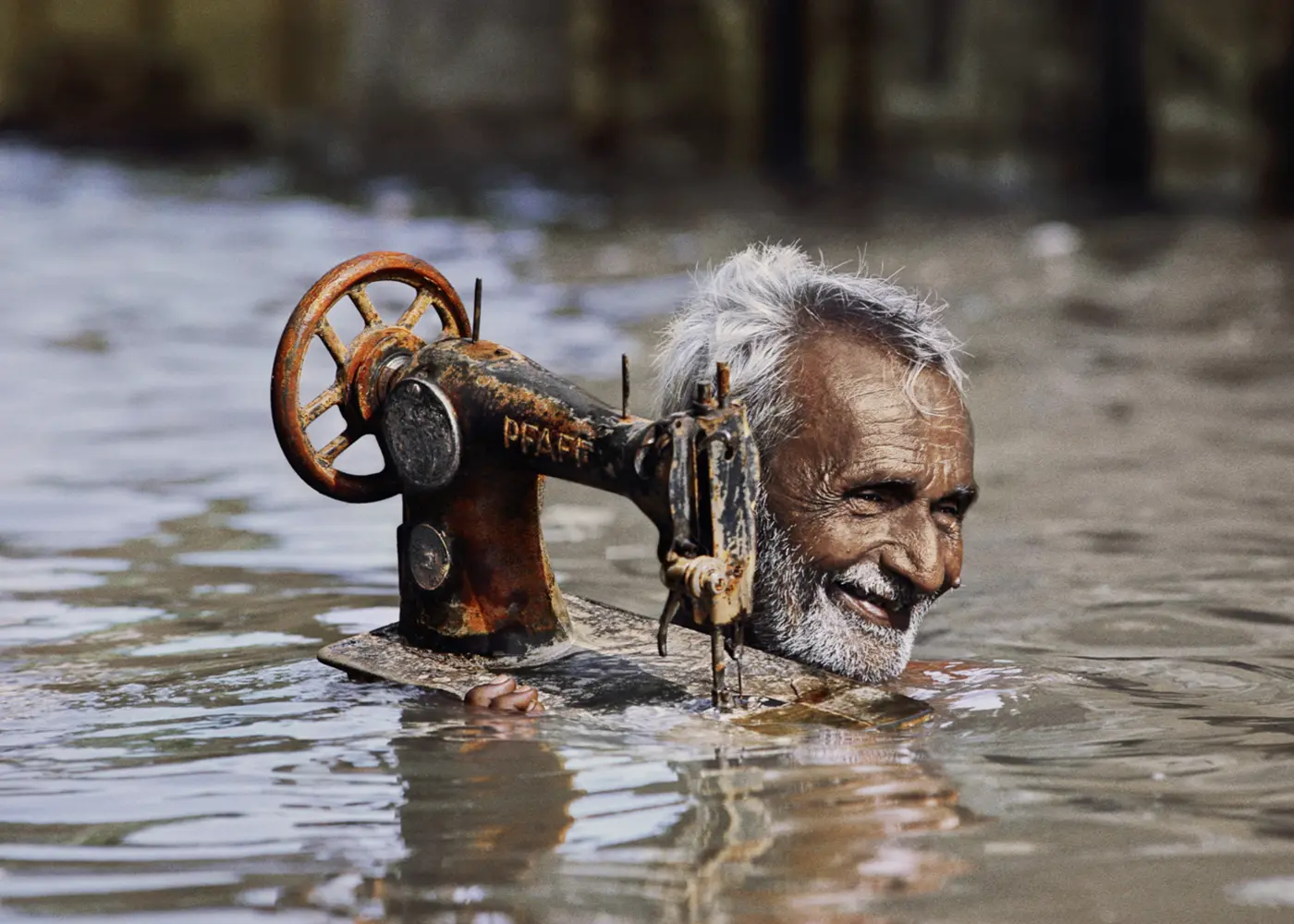 Elderly man smiling as he carries a vintage sewing machine through floodwater, with only his head and the machine visible above the surface.