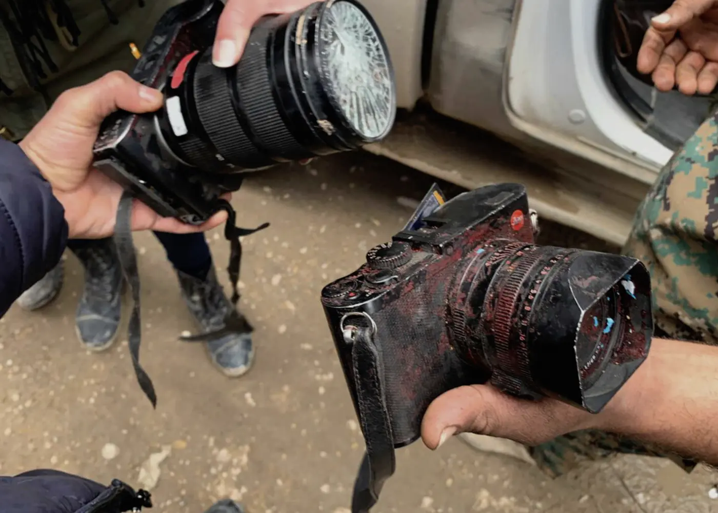 Close-up of two heavily damaged Leica cameras being held after an incident, showing a shattered front element and scratched, dirt-covered camera bodies.