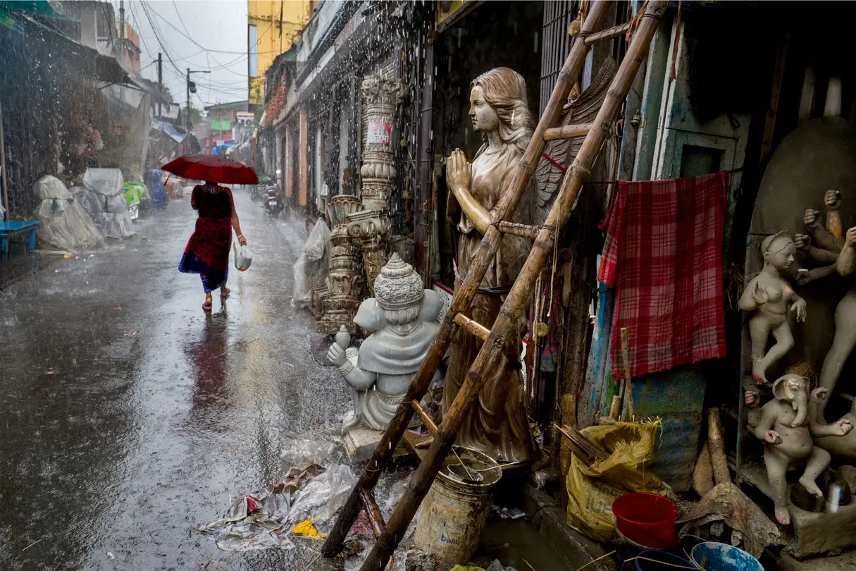 Rainy alley with statues. A woman with a red umbrella walks through a rain-soaked alley in India beside unfinished religious statues and sculptural figures.