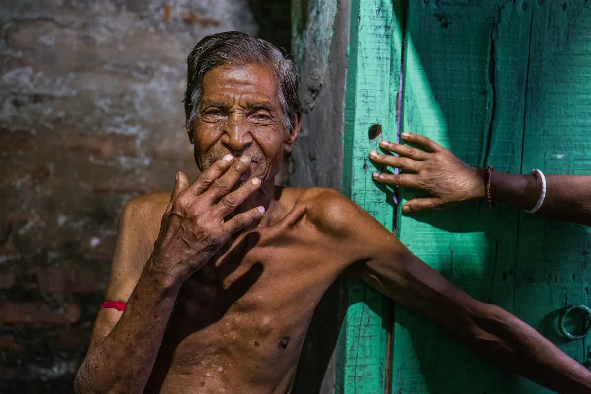Portrait at a turquoise doorway. An elderly man stands shirtless beside a turquoise wooden doorway in India, captured in strong natural light with another person’s hand entering the frame.