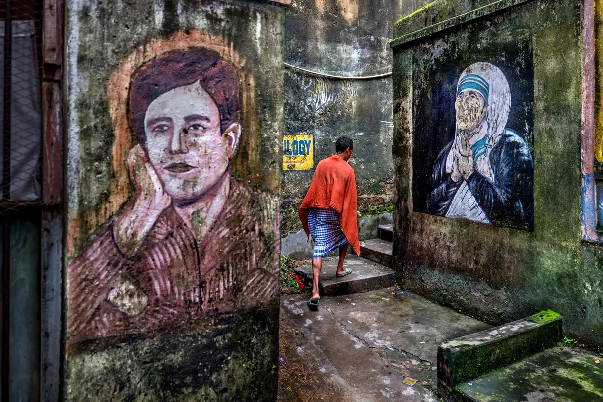 Street art and Mother Teresa mural. A man walks through a weathered alley in India between large painted murals, including a portrait of Mother Teresa on a moss-covered wall.