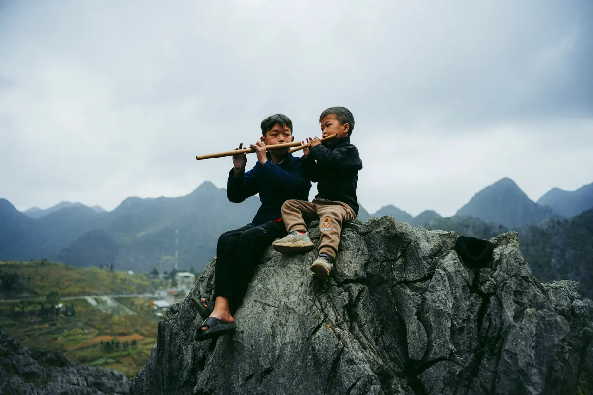 Children playing flutes, Two children sit on a rocky outcrop playing wooden flutes against a dramatic mountainous landscape.