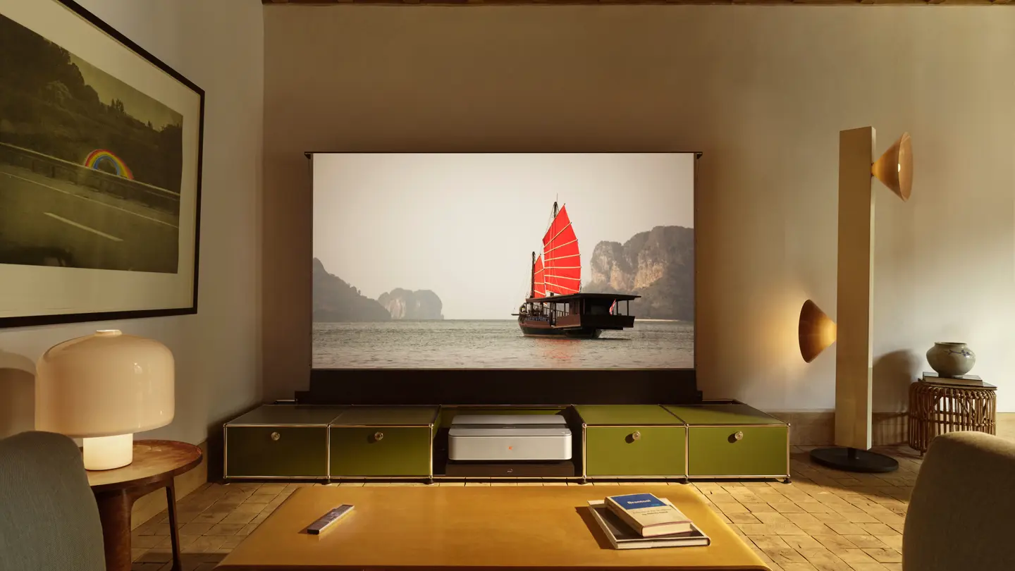 A warmly lit living room with exposed wooden beams, a sofa and a low coffee table. A large projection screen displays a traditional boat with bright red sails on the water, with a projector positioned beneath the screen.