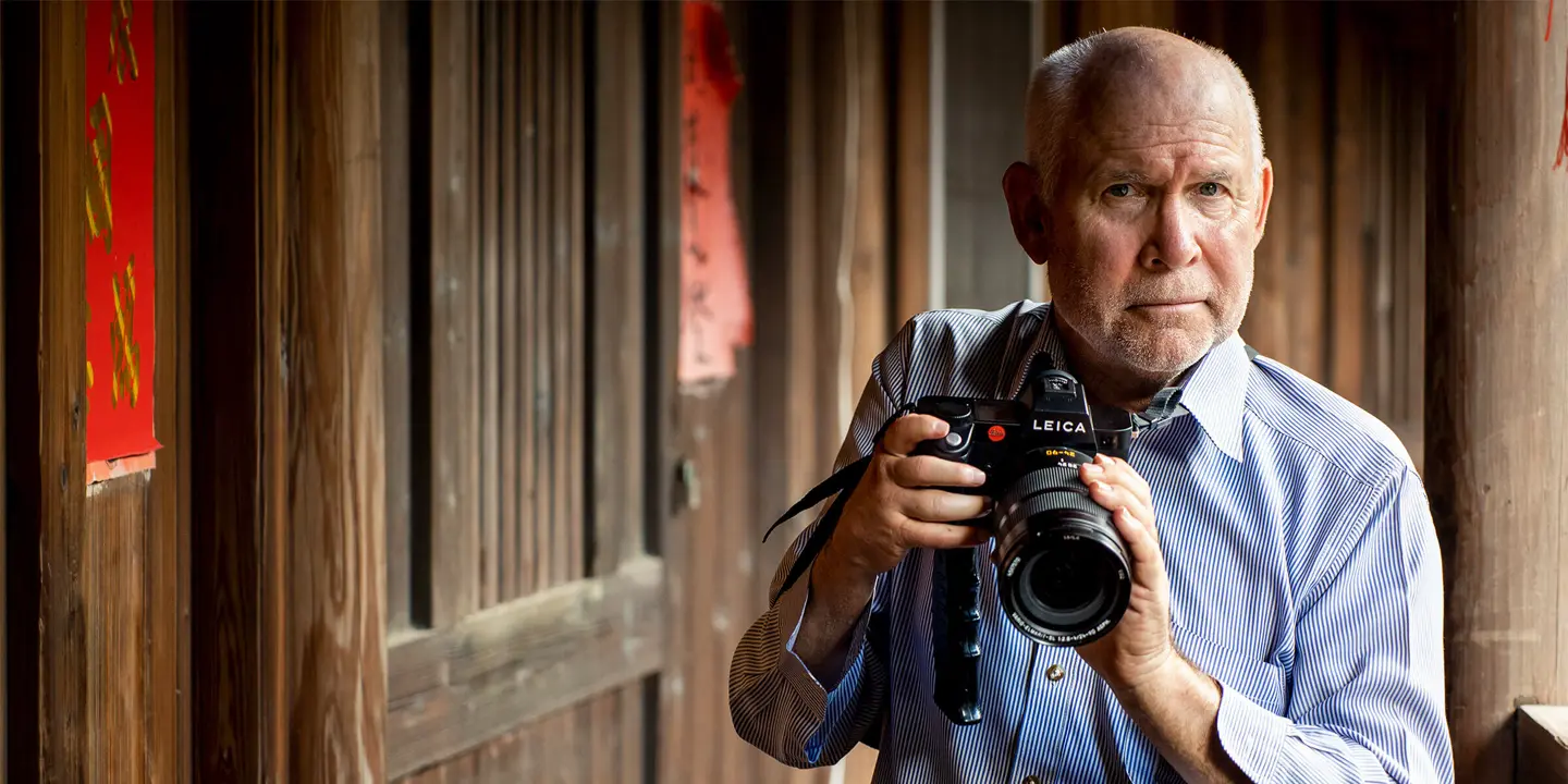 Steve McCurry holds a Leica camera and stands in front of a wooden house.