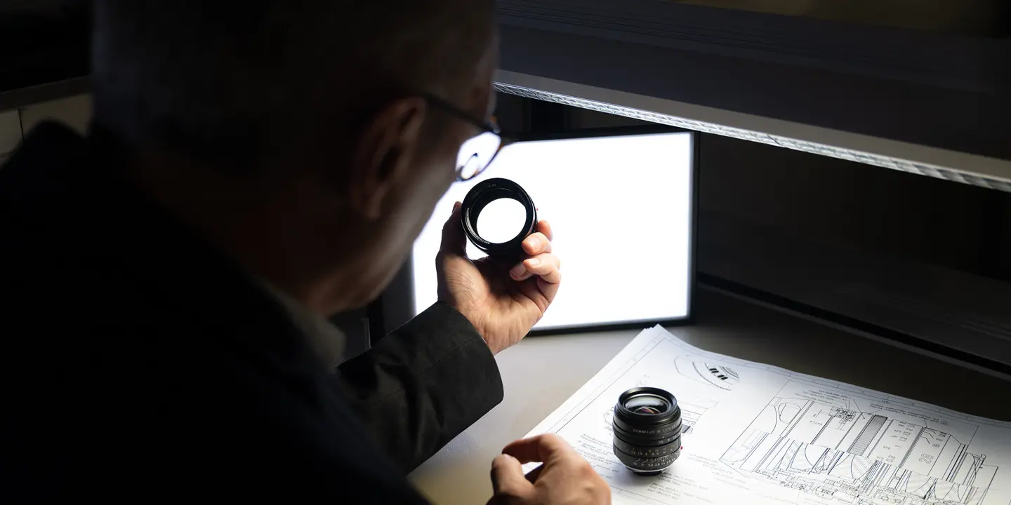 A man inspects a camera lens, technical drawings lie on the table.