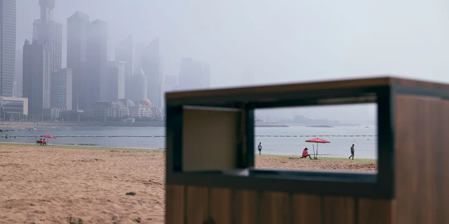 View through the rectangular opening of a garbage can on the beach with skyscrapers in the foggy background.