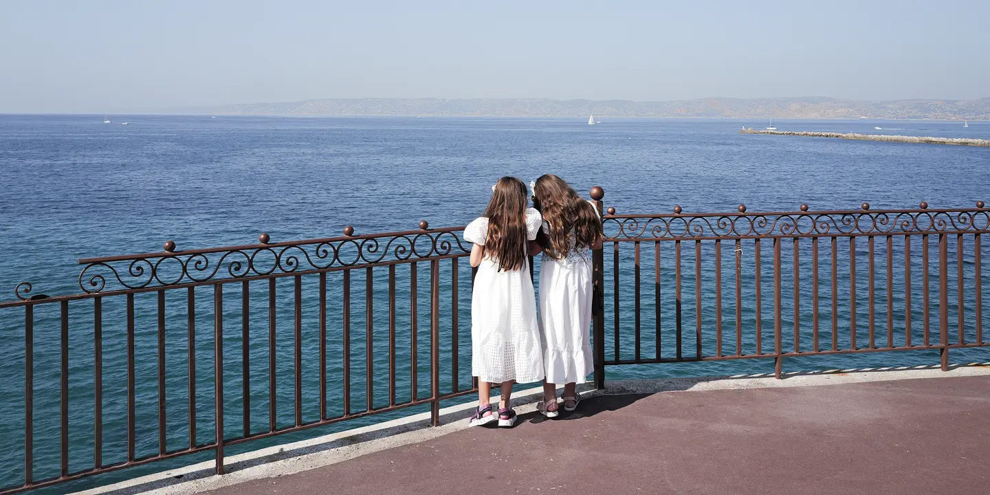 Two girls in white dresses stand at a railing and look to the sea.