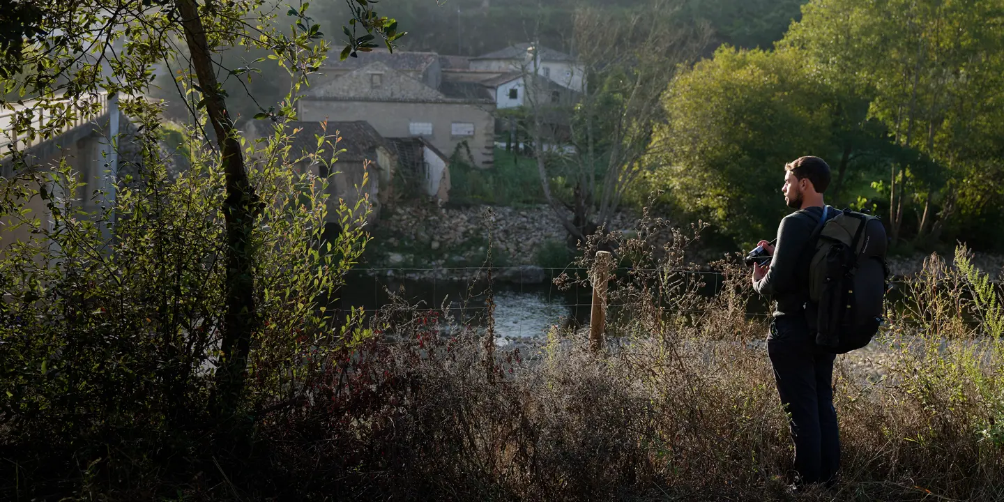 A man with a rucksack stands in the countryside and looks out over a small settlement by the river