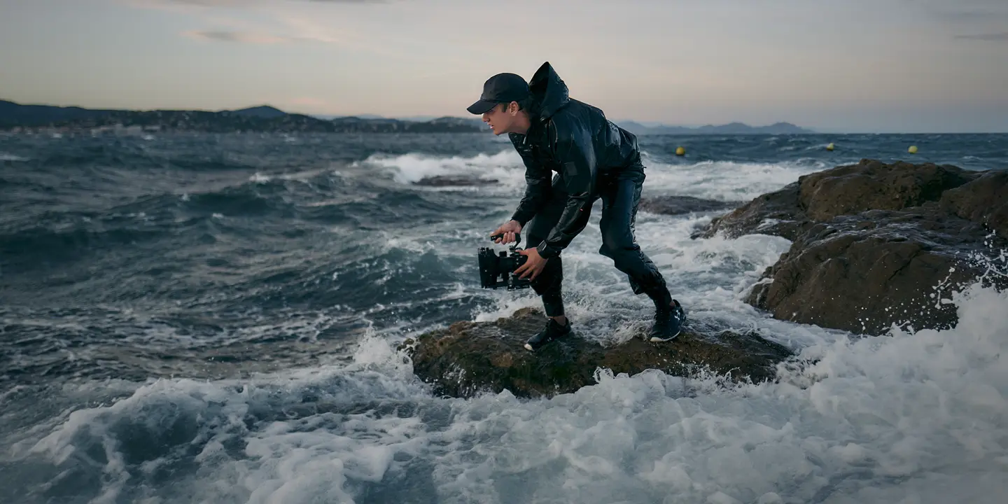 A person is standing on a rock in a stormy sea, holding a pair of binoculars.