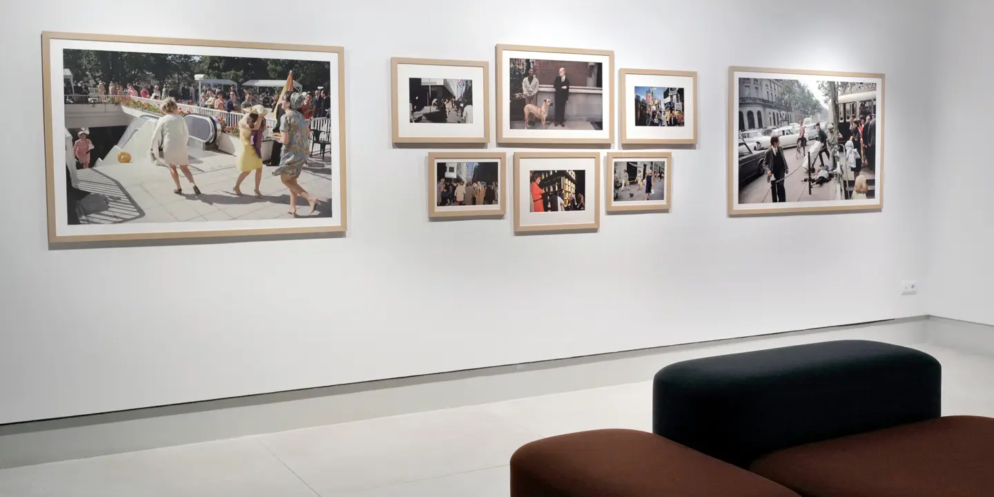 Framed pictures of people in the city hang on a white wall, with black and red upholstered stools in front of them.