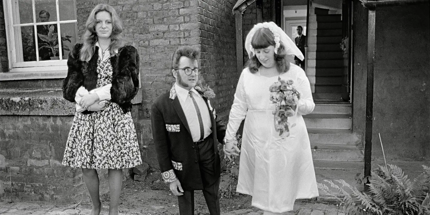 A wedding couple and a woman in a dress stand in front of a building.