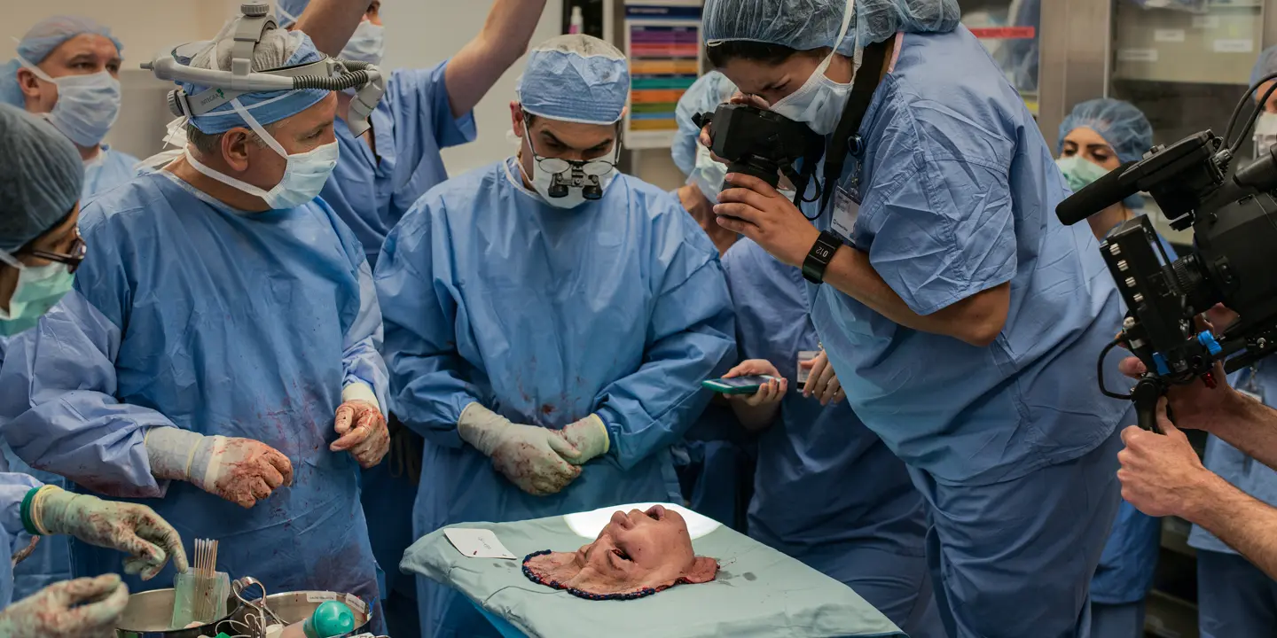 A group of people in surgical gowns look at a mask representing a face, and some take pictures of it.