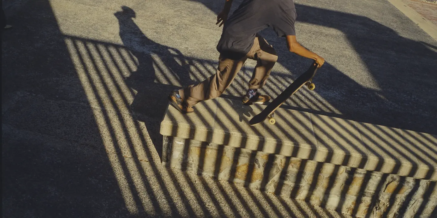 Skateboarder carrying a board through dramatic striped shadows at a skatepark, capturing movement, urban culture and graphic light.
