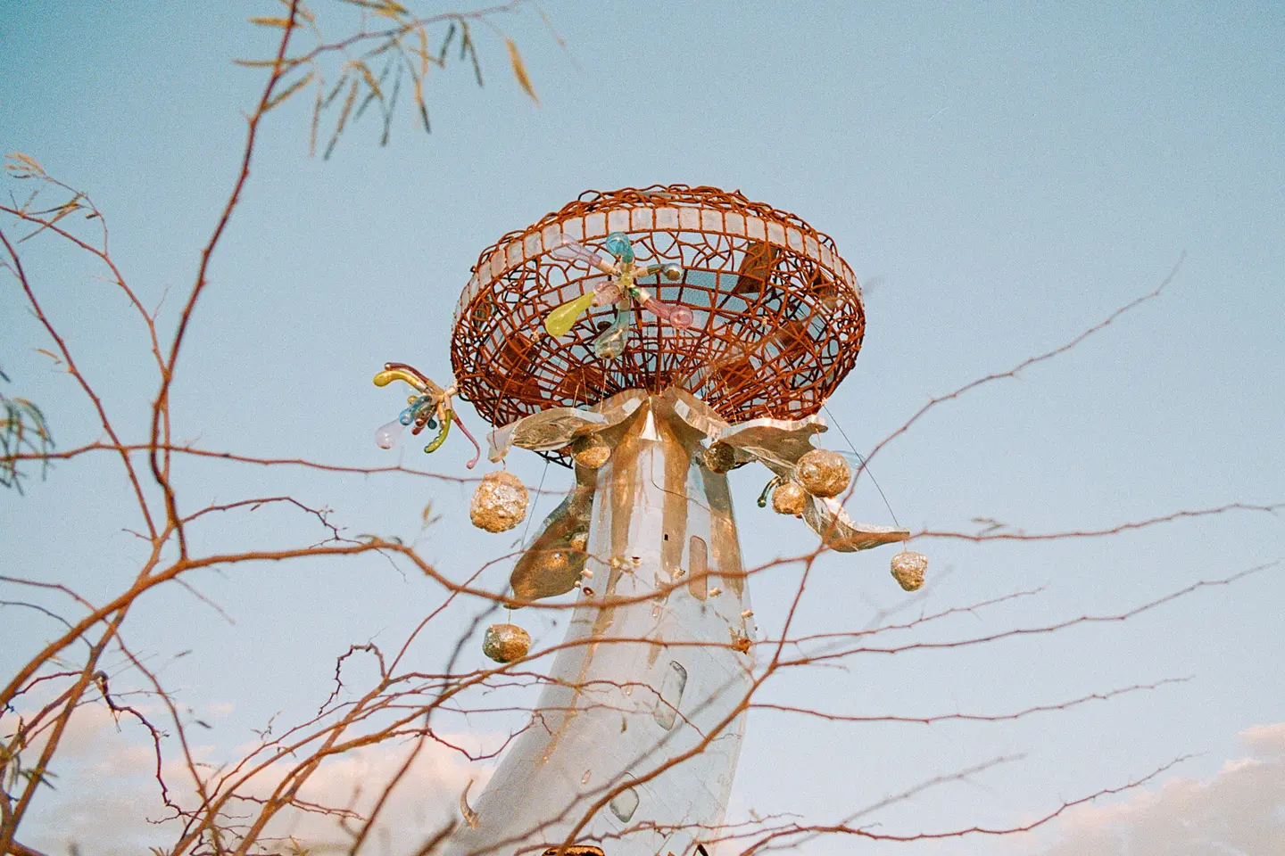 Metallic sculpture with colourful details in front of a blue sky and branches.