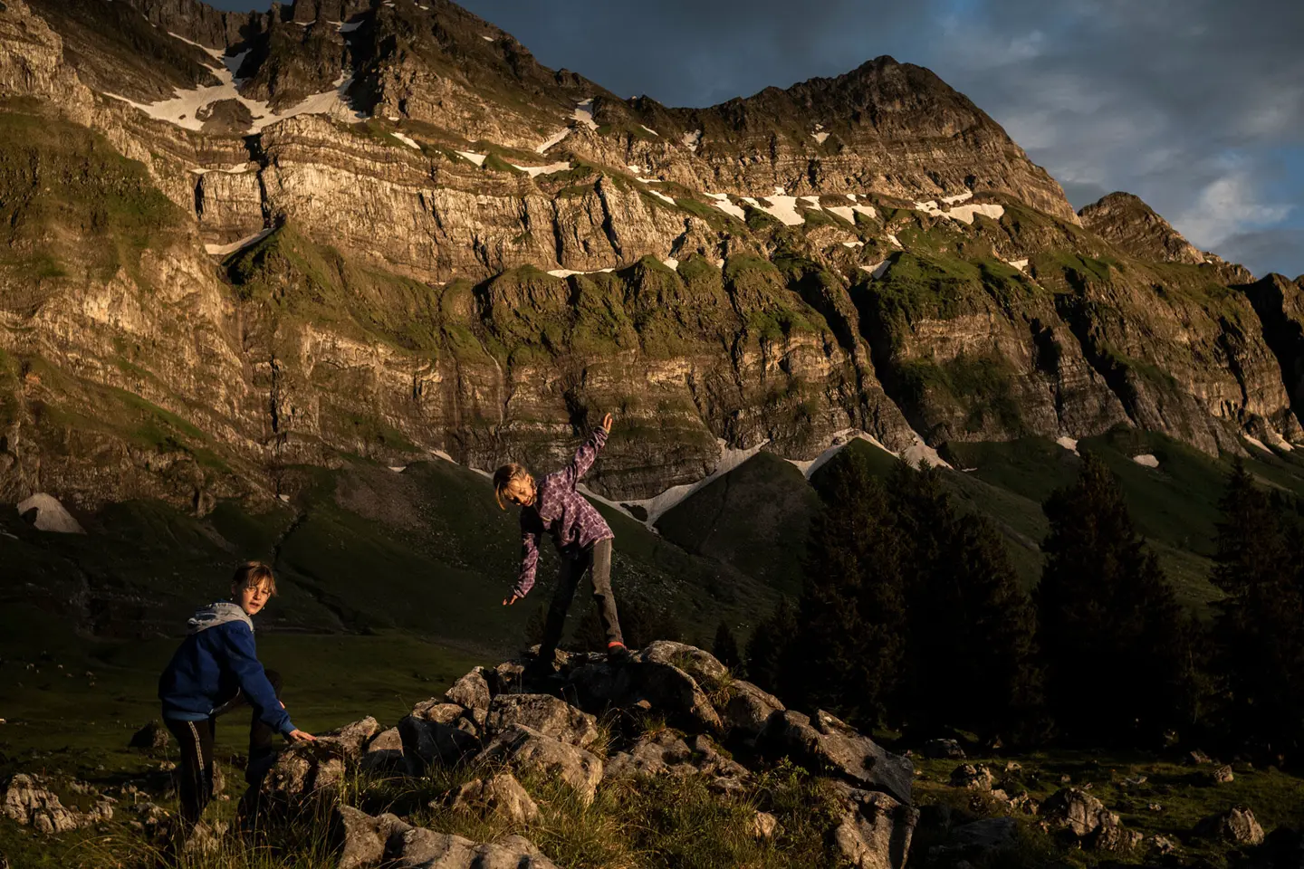Two children climbing rocks in a mountain landscape at sunset.