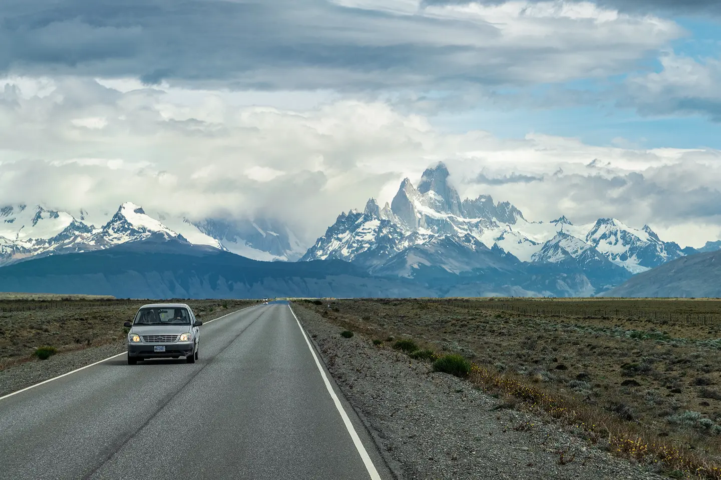 A car is driving on a road with snow-covered mountains in the background.
