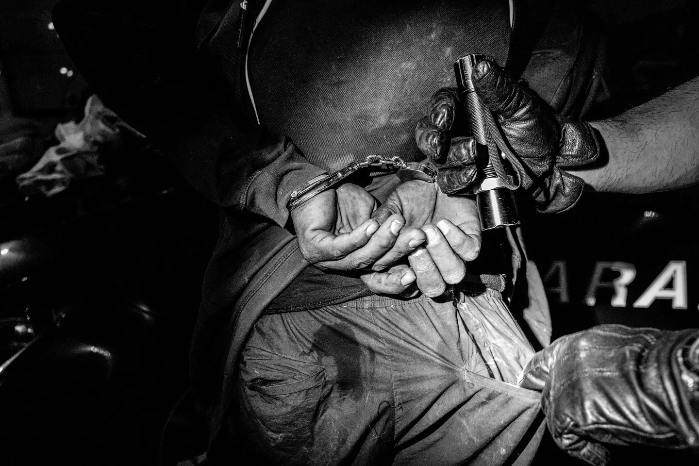 Close-up of a handcuffed person and another person searching his pants pocket with a flashlight.