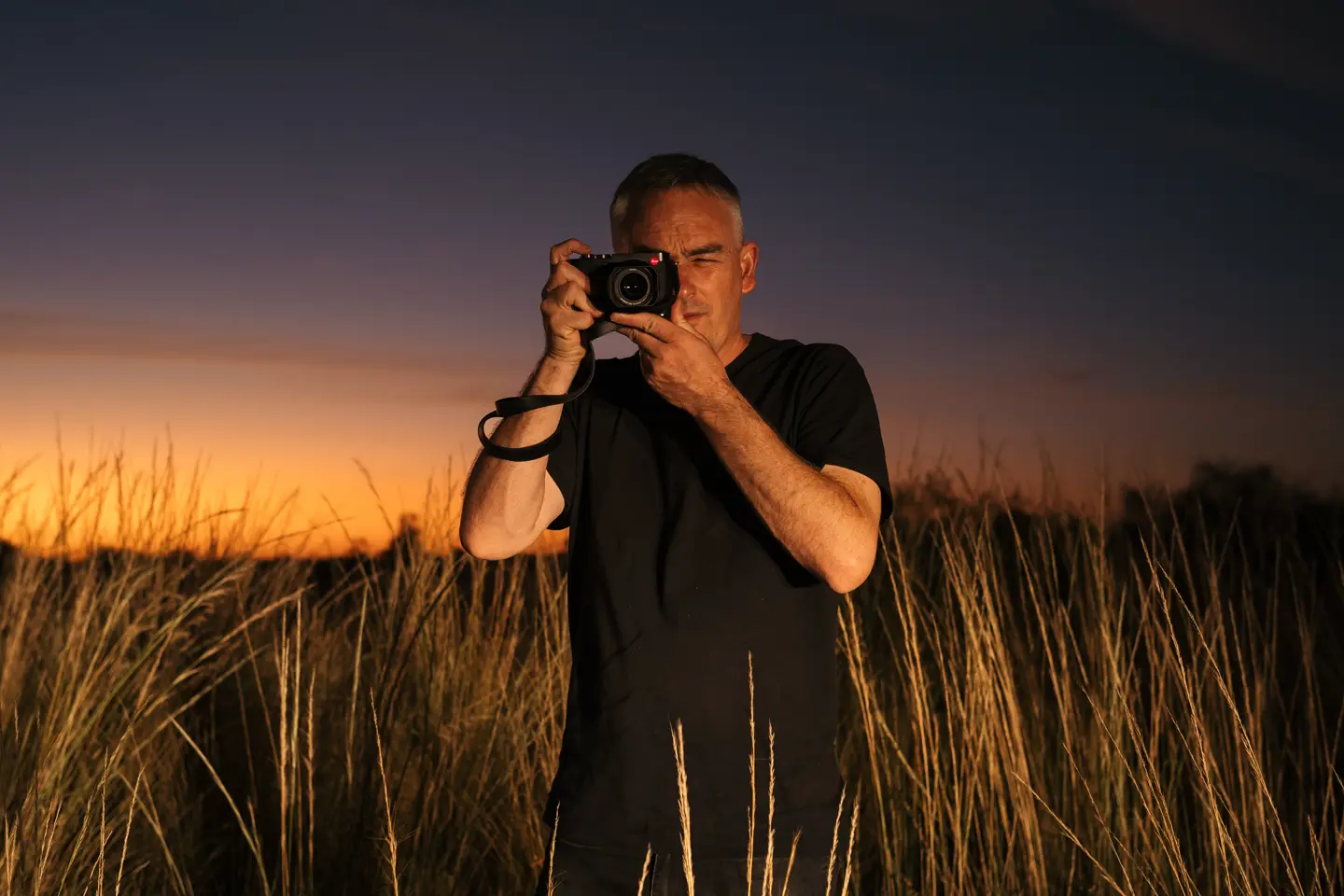 Person in a field with tall grass taking photos with a camera at sunset.